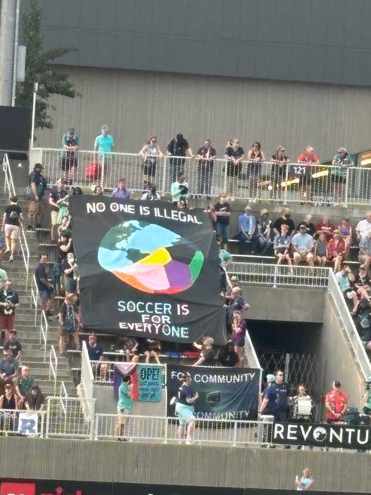 Colorful banner with the words: No On Is Illegal, Soccer is for Everyone. There is a half globe/half soccer ball in rainbow colors. A group of people are holding it above their heads at a soccer game.