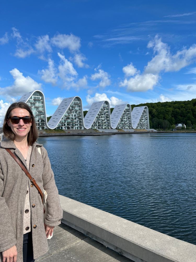 Woman smiling, standing in front of a row of five apartment buildings shaped like rolling waves, on the edge of the water, in Vejle, Denmark.