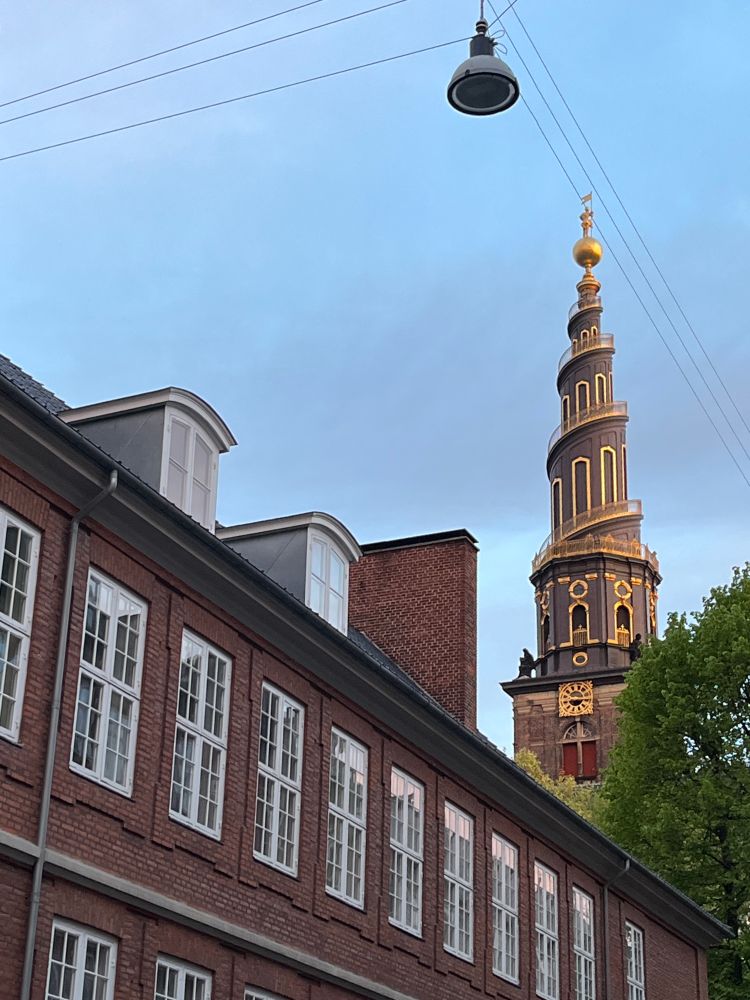 The spiral tower of Church of Our Saviour (Vor Frelsers Kirke) in Christianshavn, Copenhagen, rising above the top of an old brick building with rows of white windows. The evening sky in the background is pale blue.