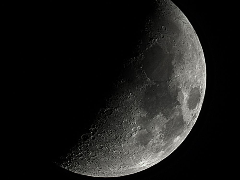 An image of the Moon in the nearly first quarter phase showing many impact craters along with smooth dark plains. 
