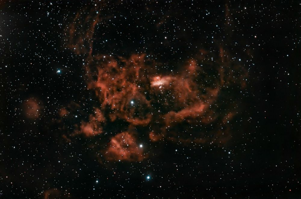 A telescopic image of stars. There's a large red nebula with an irregular splotchy shape at center. 