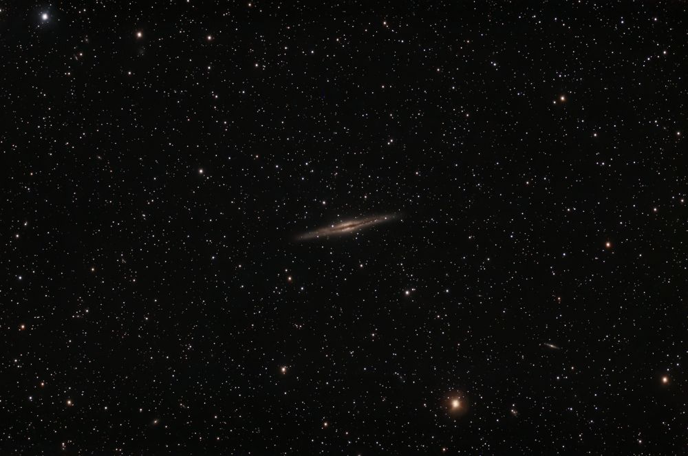 A telescopic image of stars. At center is a spiral galaxy seen from the side. It's elongated with a bulge in the center. A dark lane of dust cuts along the length of the galaxy.  A second smaller looking galaxy can be seen to the upper right of the orange star near the bottom of the image.