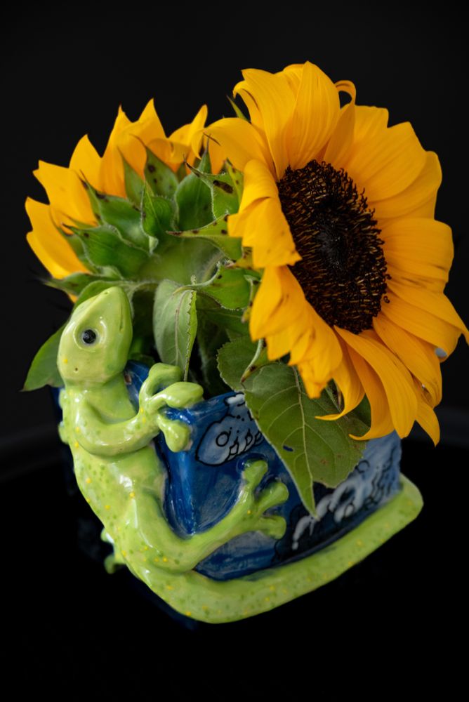 A ceramic flower pot filled with sunflowers. The ceramic pot is blue and light green. It features a green gecko climbing up the side, looking over the top rim with the tail wrapping around the base of the pot. It has black beady eyes and thick suction pad toes. The blue vase is covered in white billowing cloud illustrations. The pot is sculpted with angel white #182 clay. It is painted with Mayco underglazes, black designer liner, and clear glaze. It was sculpted by Justin Groom. #ceramics #sculpture #ClayArt #pottery #art #gecko #clouds #sunflowers