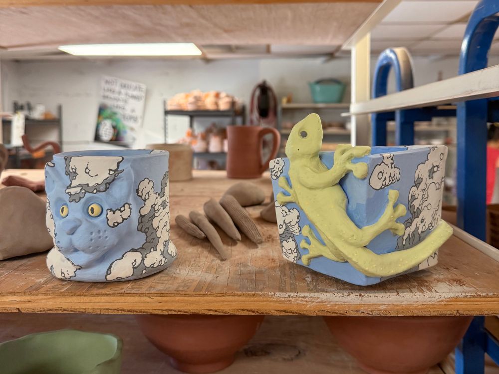 Two brightly colored ceramic pieces are sitting on a shelf, waiting to be bisque fired. One is of a coffee mug, with a cat face and tail as the handle the other is of a flower pot with a gecko lizard on it both are painted with clouds and blue sky.