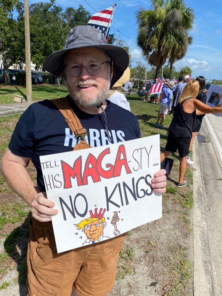 A bearded man in a grey sun hat, a Harris Walz shirt, glasses, and brown overall shorts is holding a protest sign. The sign reads: tell his MAGA-sty no kings. It has a drawing of trump with a shit crown and scepter. It’s a very sunny day. The protester is smiling from all the people and supporters.