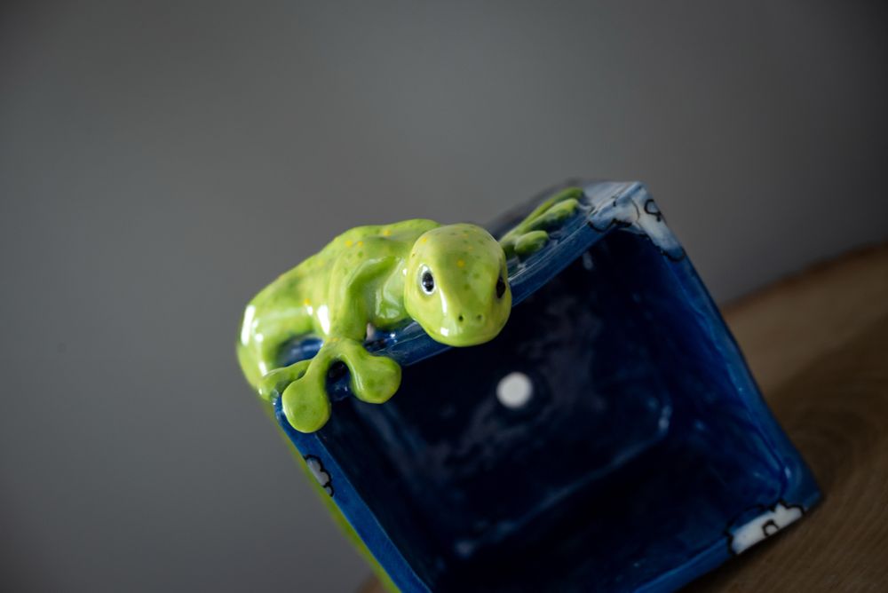A gecko flower pot. A green gecko with black eyes is crawling around a square flower pot. His head is peaking over the top of the pot. The pot is blue with illustrated white and gray billowing clouds. The entire sculpture is glossy and shiny. This picture is a top view looking down.

#ceramics #ClayArt #pottery #plants #gecko #sculpture #ArtTherapy #art