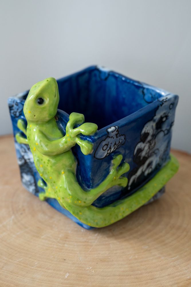 A gecko flower pot. A green gecko with black eyes is crawling around a square flower pot. His head is peaking over the top of the pot. The pot is blue with illustrated white and gray billowing clouds. The entire sculpture is glossy and shiny.

#ceramics #ClayArt #pottery #plants #gecko #sculpture #ArtTherapy #art
