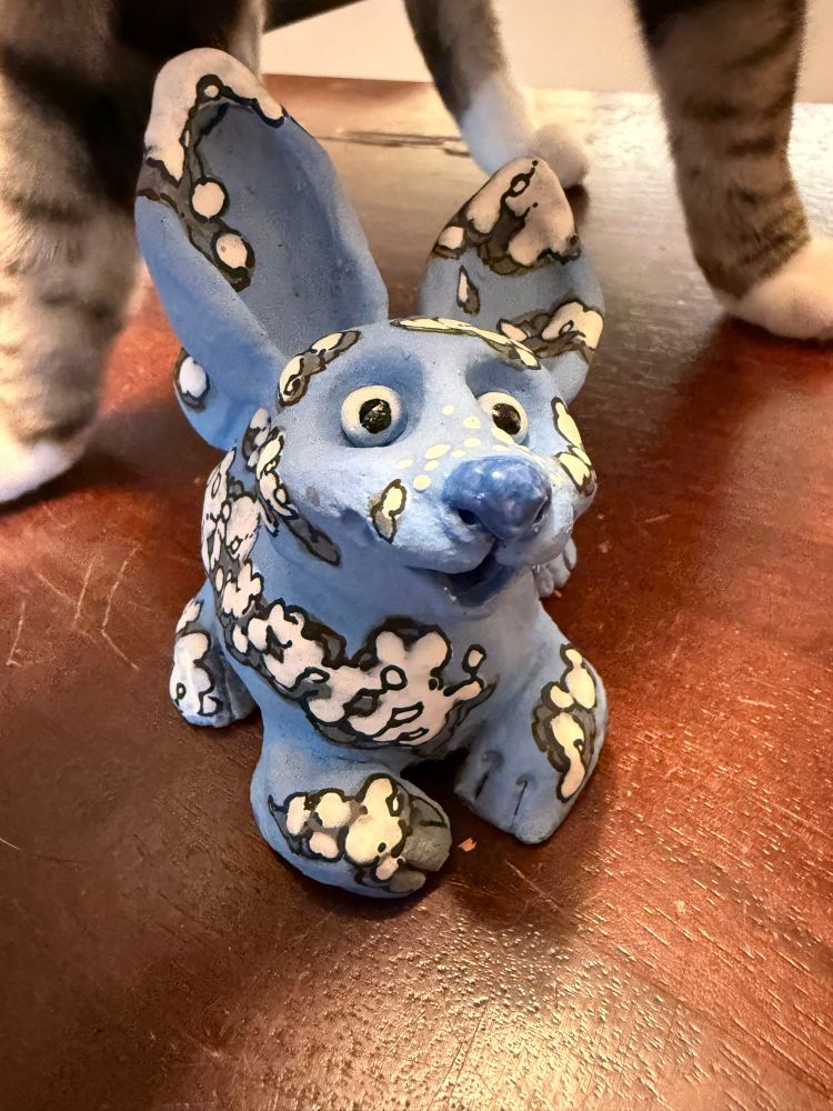 A #whimsical and #cute #bunny #sculpture is looking up at the camera. It is blue with white billowing #clouds. It’s on a dark wooden table with a #tabby cat’s feet walking in the background.

#ceramics