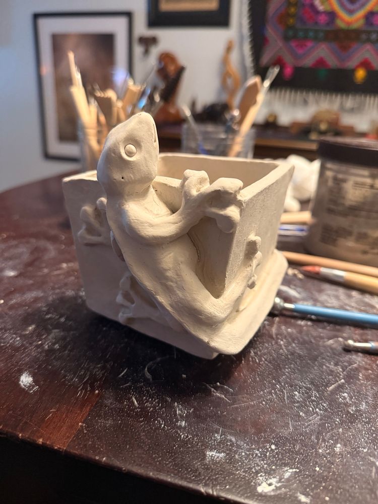 An off white clay sculpture sits on a dark wooden table and is surrounded by sculpture tools. The sculpture is of a gecko lizard climbing around a square flower vase.