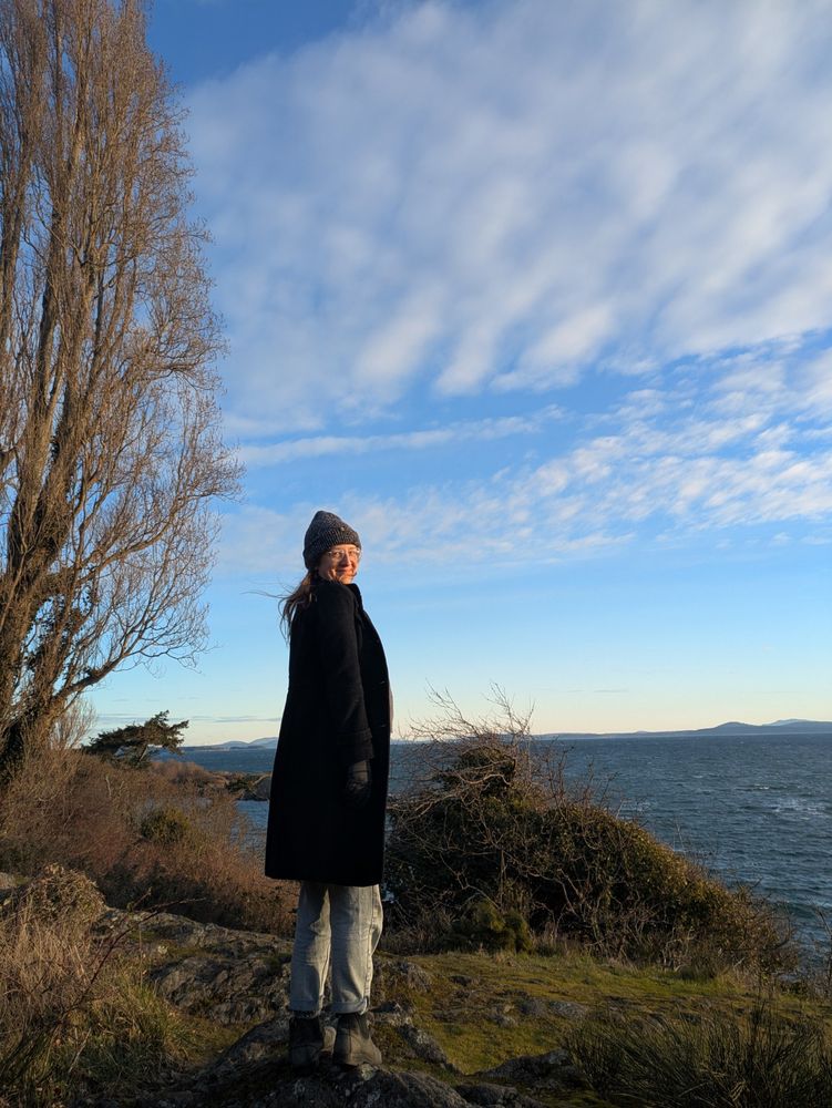 Woman in a black coat and grey toque smiles beside the Pacific Ocean 
