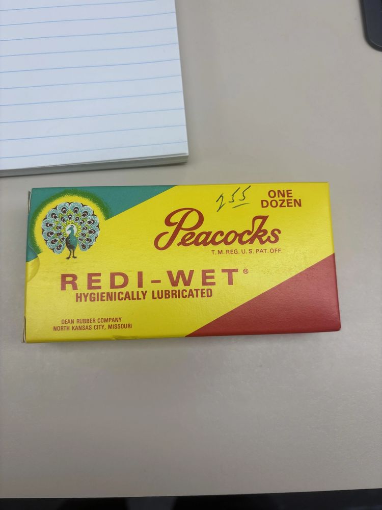 Photo of an antique condom box. The box reads " One Dozen; Peacocks; Redi-Wet; Hygienically Lubricated; Dean Rubber Company; North Kansas City, Missouri". The box is teal, yellow, and red striped with an illustration of a peacock in the upper left corner