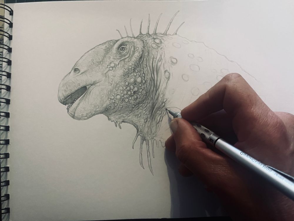 A photograph shows my hand holding a pencil, working on a portrait of a long-necked dinosaur with wrinkled, knobbly skin. 