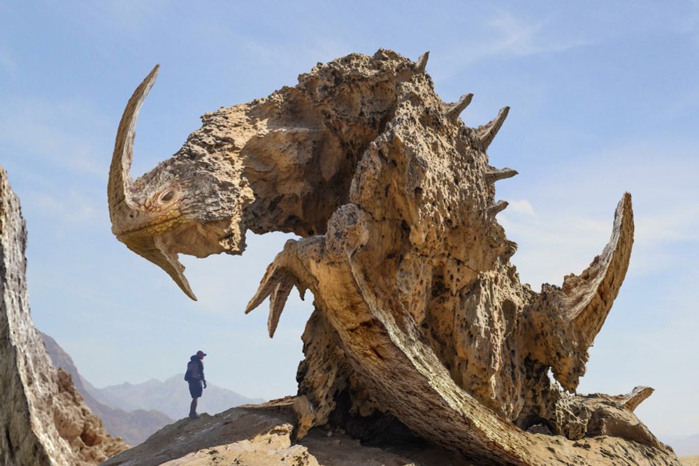 Digital illustration. A man in hiking gear stands on sandstone rocks. Looming over him is a giant rock formation, looking like a huge dragon, with horns on its head and spikes along its back. It appears motionless, froze in time. 