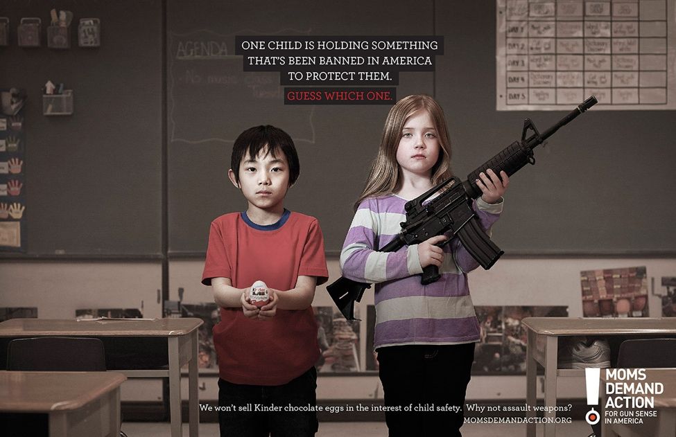 Boy holds a Kinder Egg in a classroom. A girl holds an automatic firearm. One is illegal. The Kinder Egg is illegal in the United States.