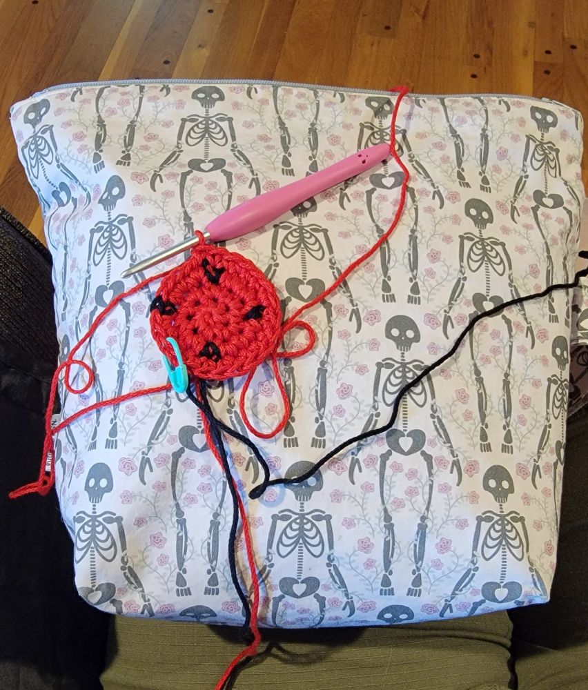 The beginning center of a watermelon kippa rests on a yarn bag decorated with skeletons and flowers
