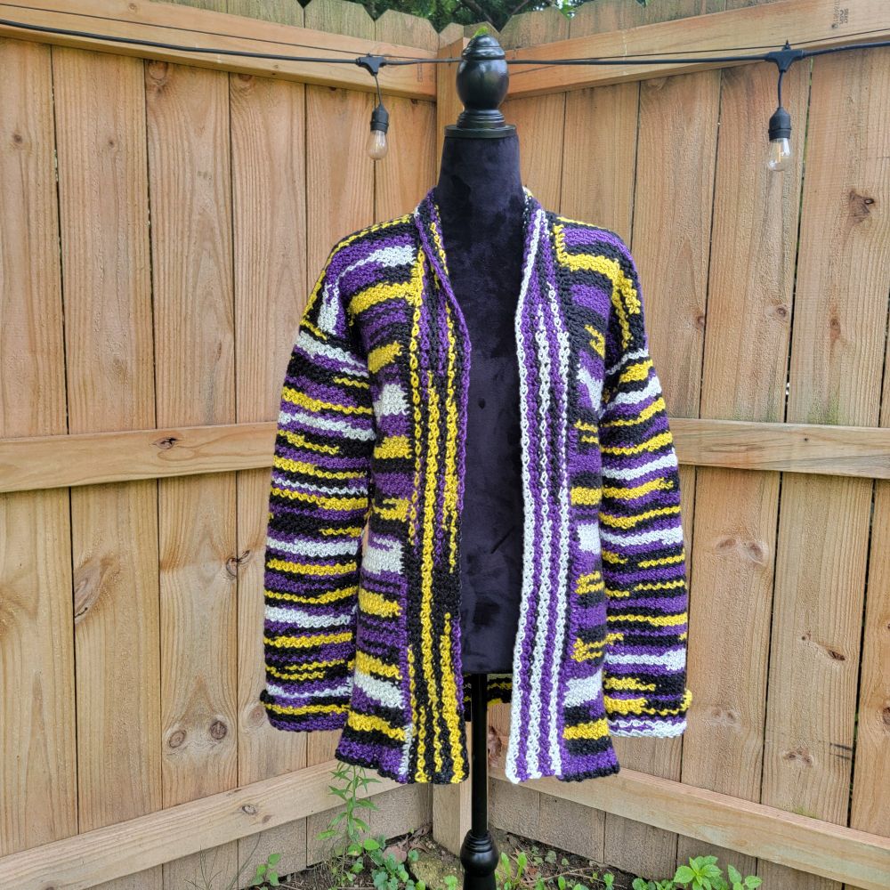 An open cardigan sweater in nonbinary pride colors hangs on a black dress form in front of a blonde wooden fence