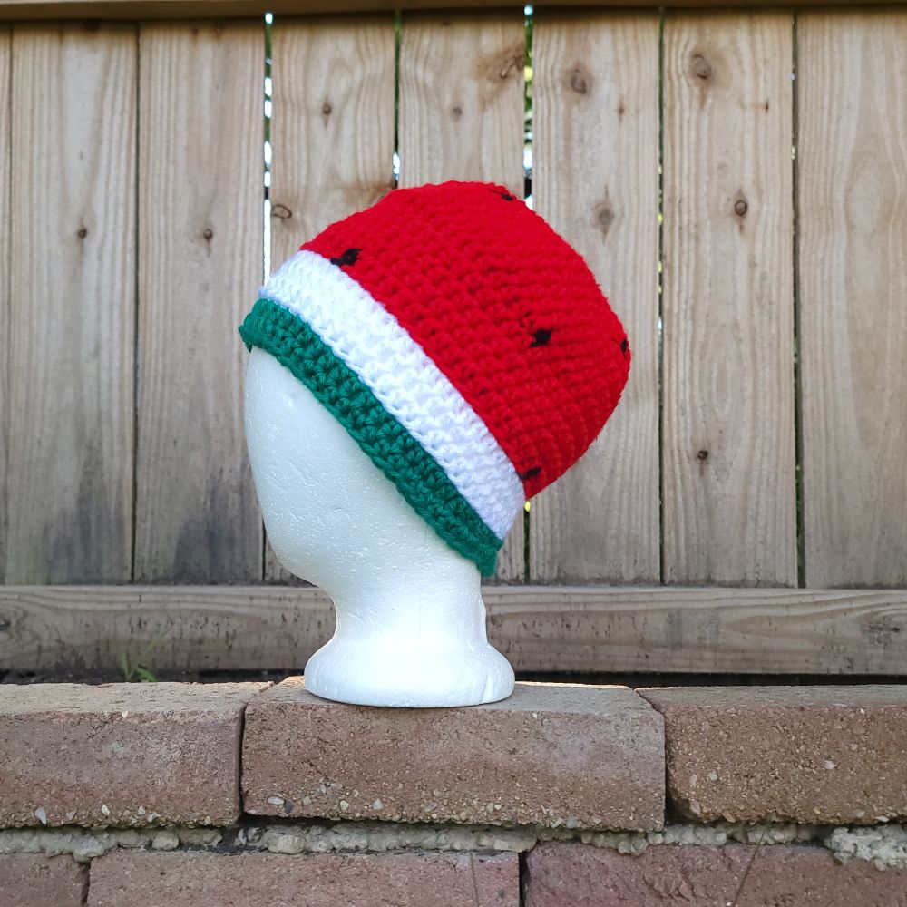 A crocheted beanie had made to look like a watermelon cut in half rests atop a foam head sitting on a brick wall, a wooden fence in the background 
