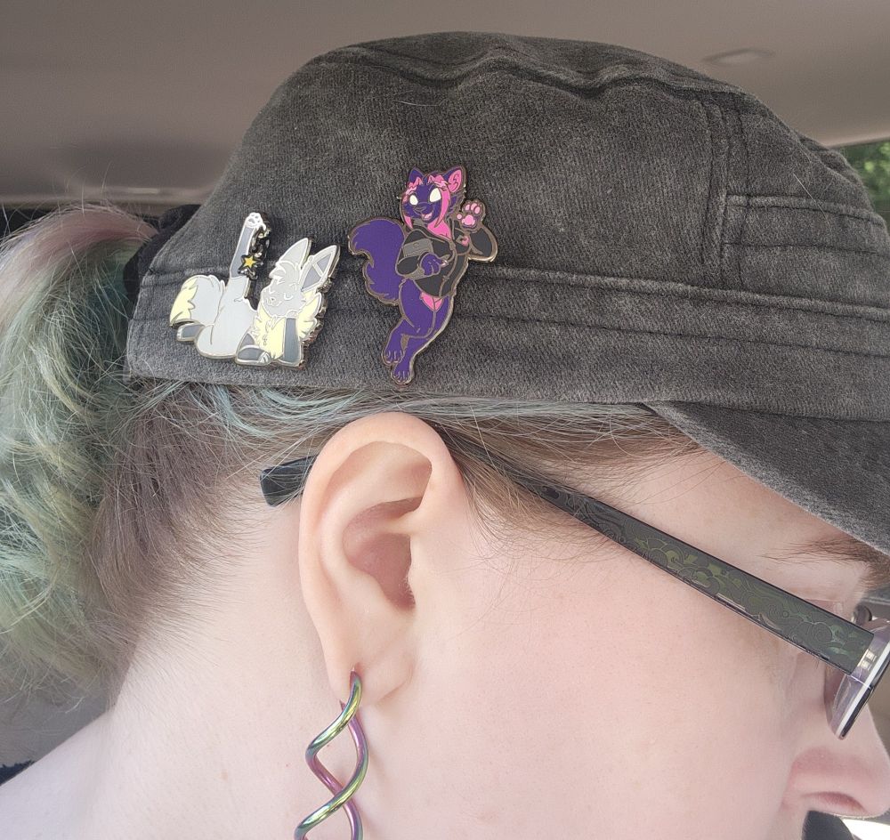 Picture showing two enamel pins on my hat, one is my purple and pink cat character and the other is a Shiny eevee, representing my late friend Dragoneer