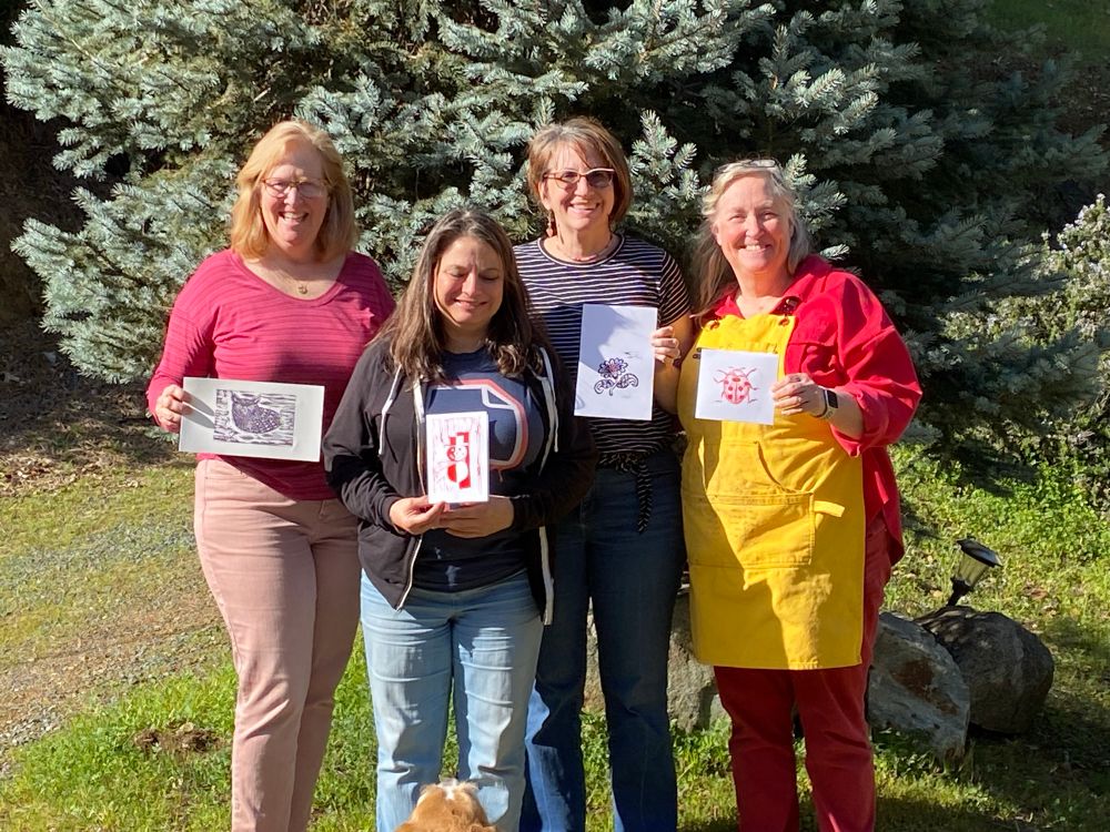 Four smiling friends holding their printed linocuts creations outside. Three had just spent hours learning about how to carve a linocut block and then print it. The teacher is the woman in the yellow apron holding a print of a ladybug she created as an example for the class. The other prints include an owl, snowman and flower. 