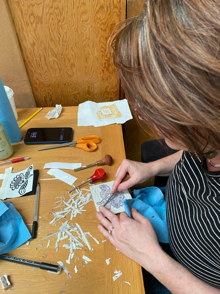 A woman cutting into a linocut block, slowly carving out an image of a flower. Shavings and tools litter the table around her. 