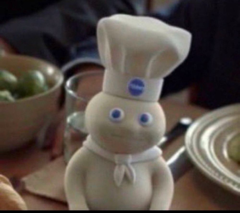Image of the Pillsbury Doughboy on a table with food, drink, and a place setting behind him. It's a still from a commercial, and it looks like he is looking sideways up at the camera with a 😐 expression 