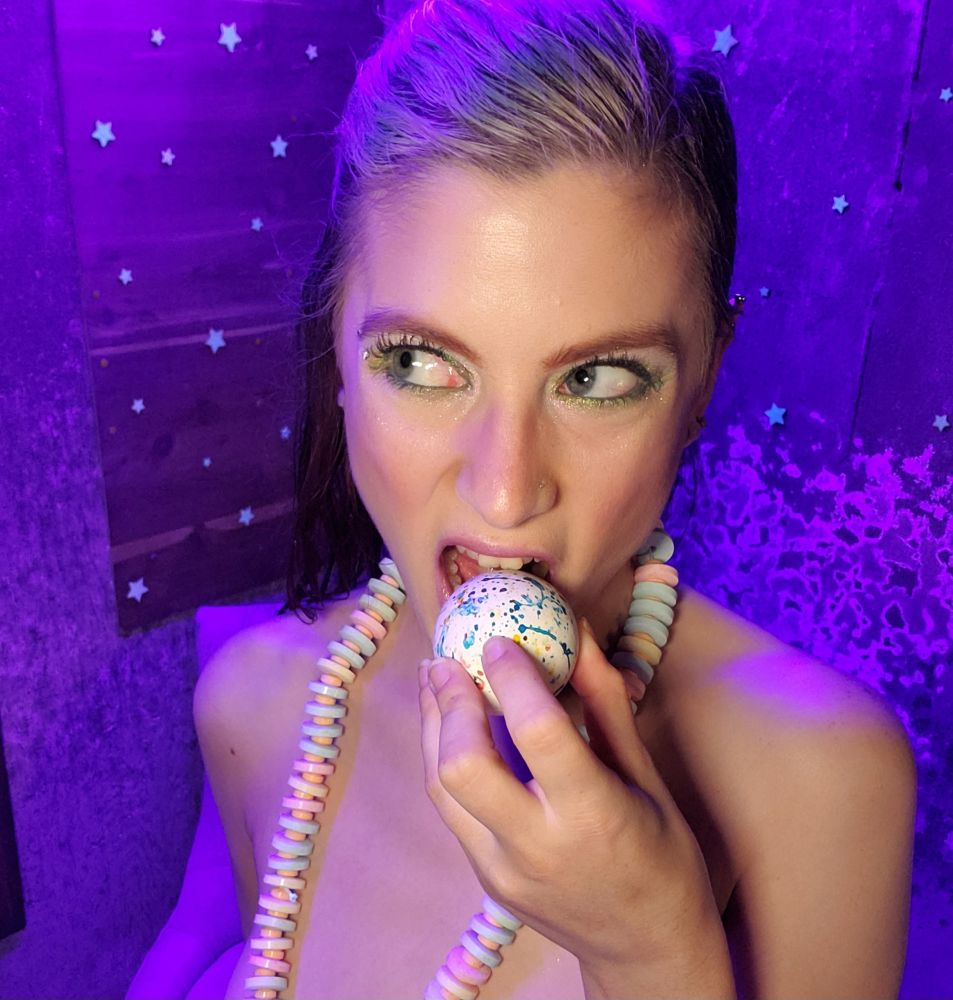 Nox standing in front of a wall of stars, wearing only a candy necklace. They are looking to the left as they bit a huge jawbreaker 