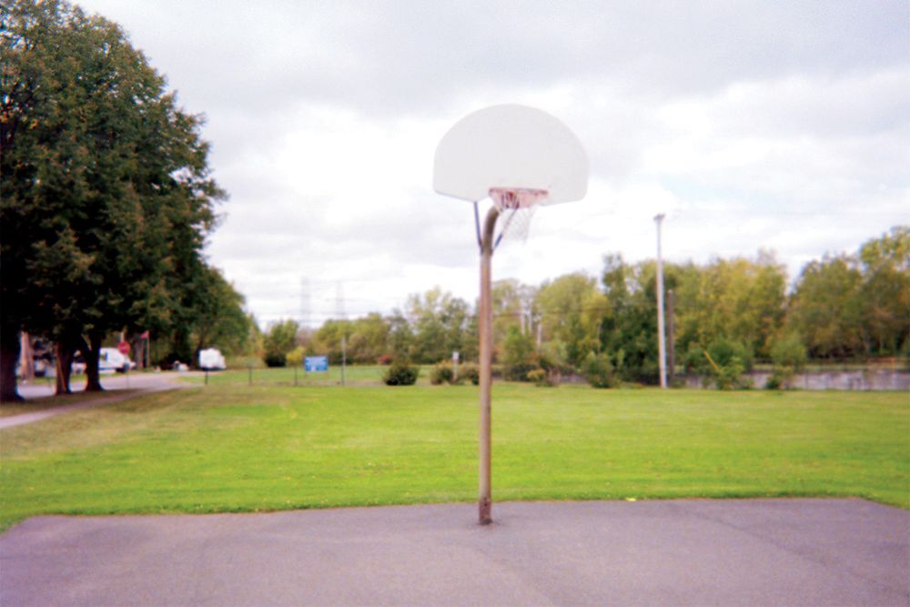 A lone basketball hoop on an asphalt court in a small city with grass and trees around it.