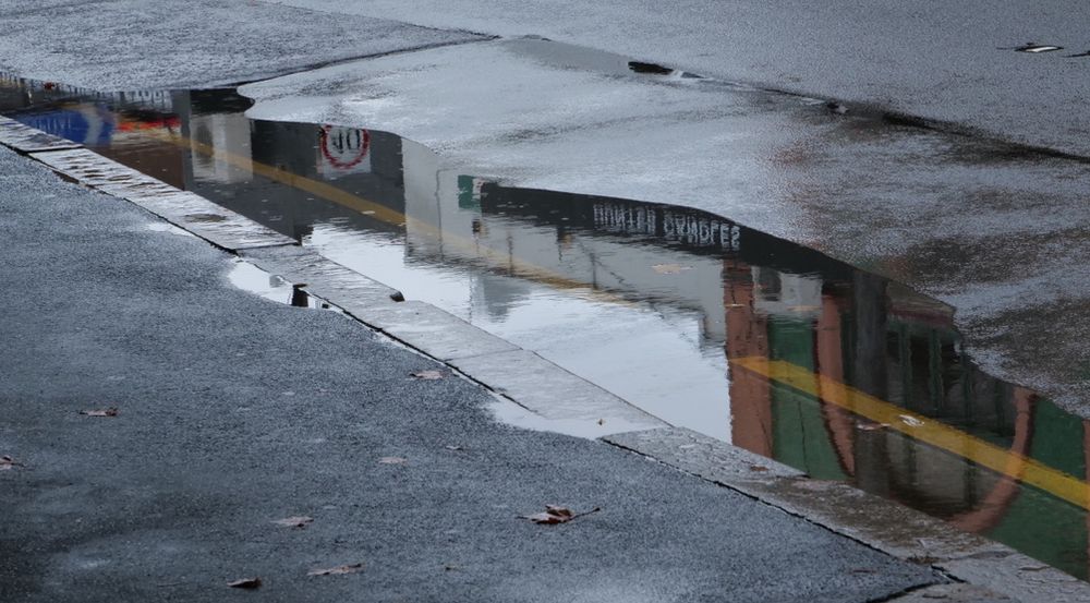A puddle on a wet road running diagonally through the middle of the frame, with footpath on the lower left side and road on the upper right. The puddle has a reflection of shopfronts along the road, 40 km/h road sign and a blue road sign indicating a hospital zone.