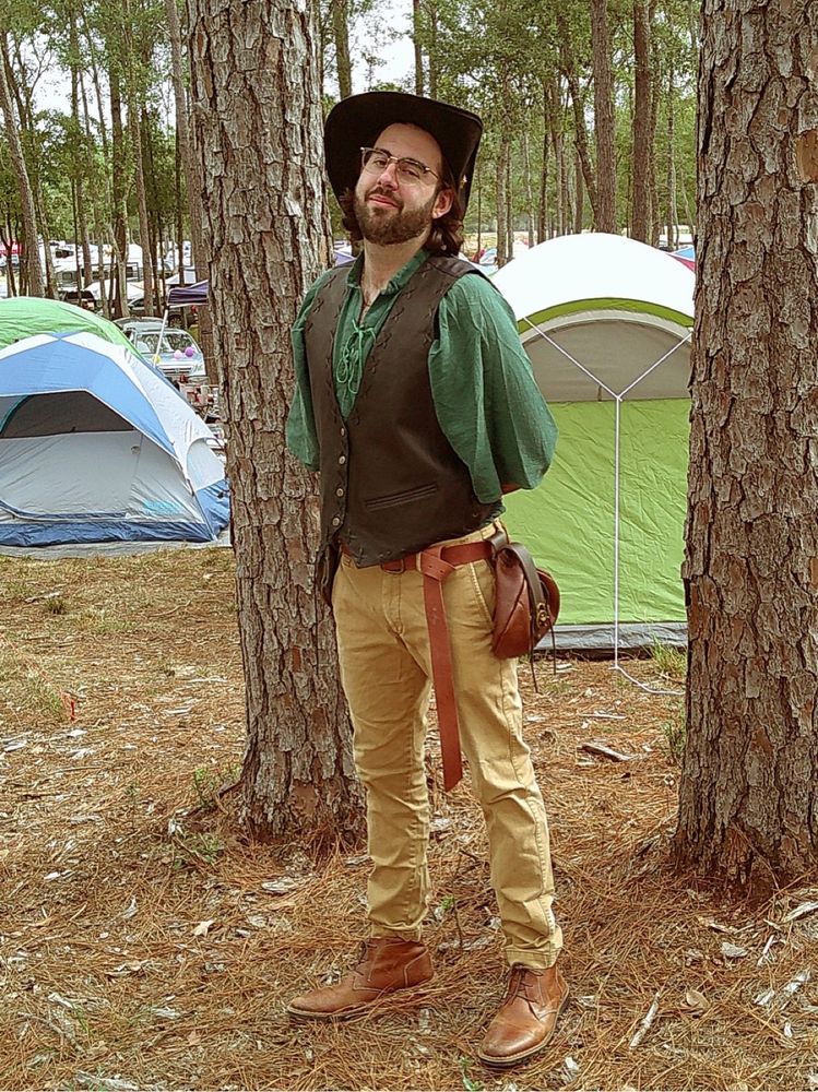 Alex in a green shirt, black vest, and tan pants, standing in a wooded campground. 