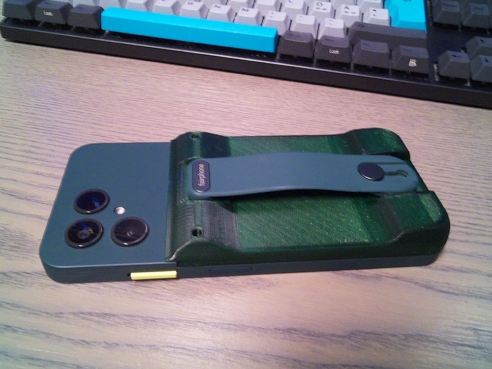 Green fairphone 6 with a 3D printed back cover. The cover is much thicker than the original one, and has a bump in both ends. The optional finger loop accessory is mounted on the new cover.