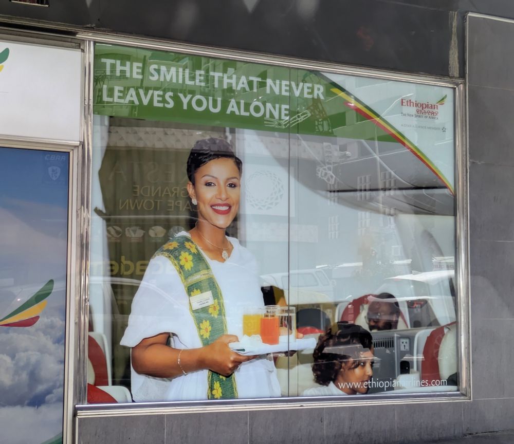 An ad for Ethiopian Airlines with a smiling woman carrying a tray making eye contact with the viewer. It reads "the smile that never leaves you alone" in all caps.
