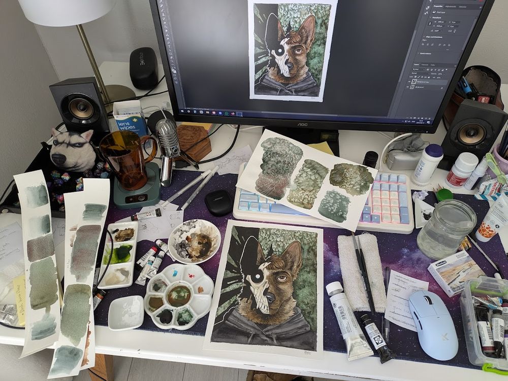 My workspace with watercolour palettes filled with browns, greens, umbers, greys, black and white. Tubes of paint are scattered around and the painting is visible in the middle on top of my desk mat. There is clutter like empty coffee cup, water jar, speakers, and art supplies.