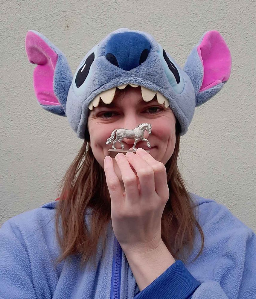 Photo of a women with long brown hair wearing a blue jacket and blue Disney Stitch hat, holding up a small metal pony in a trotting pose.