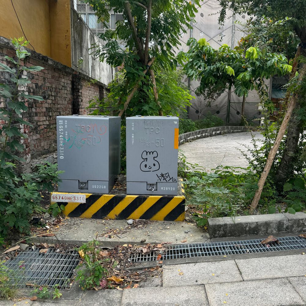 on a dark grey electricity box there is a graffiti of a character (round face with a little fuzzy top, a bit like a sprout) made of simple black lines; the electricity box is on a stand with yellow and black stripes; the box appears to be on the corner of a small park with lush green trees and weeds