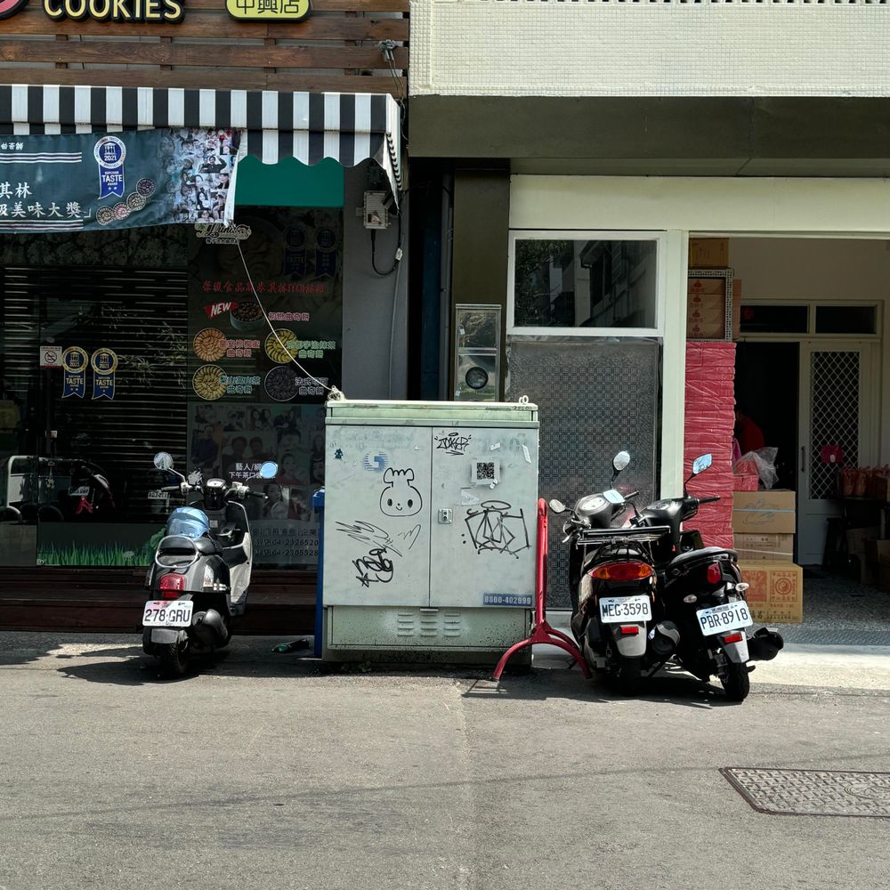 a photo taken from across the street; a graffiti of a character (round face with a little fuzzy top, a bit like a sprout) on what looks like a silver electricity box; there are two bikes on the right and one bike on the left parked; the electricity box is on the side of the street, with shop fronts behind it; there are some other squiggles and wear and tear on the electricity box