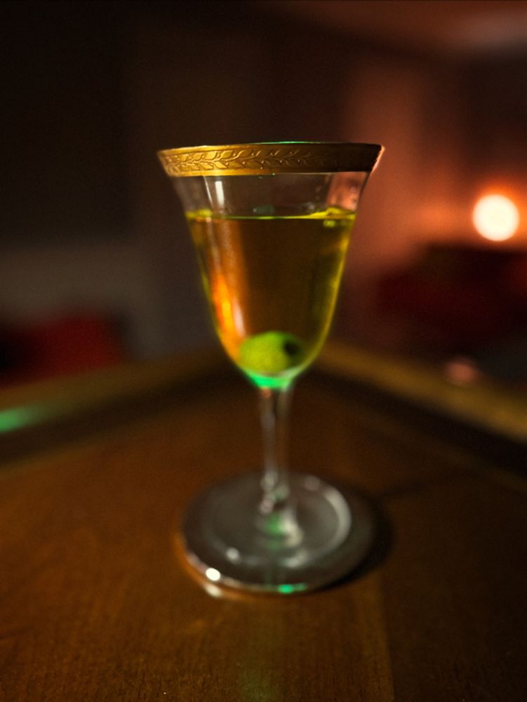 A stemmed glass with a hammered gold rim with a leaf pattern is sitting on a metallic gold coaster and resting on a wooden bar. Inside the glass there is a transparent yellow liquid with a single green olive resting on the bottom. 