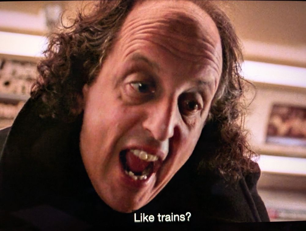 a screenshot from the movie Ghost (1990). it is a close up shot of the subway ghost character. he looks like he is speaking aggressively and the subtitle reads "Like trains?"