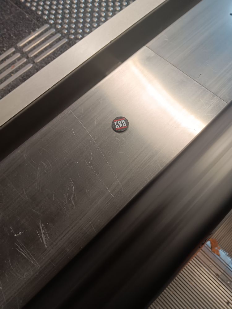 Es ist der Griff einer Rolltreppe zu sehen. Darauf ein schwarzer Einkaufswagenchip mit der Aufschrift "fck afd".