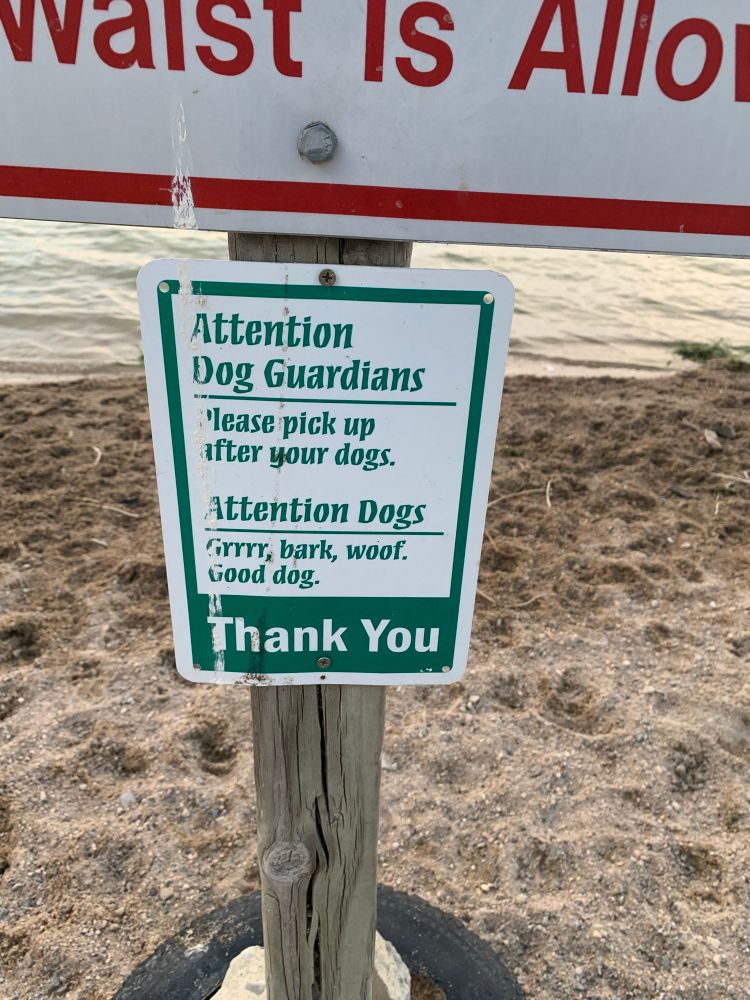 A white sign with green text. It says attention dog guardians. Please pick up after your dogs. Attention dogs, grrrr, bark woof, good dog. At the bottom and white text on a green background it says thank you