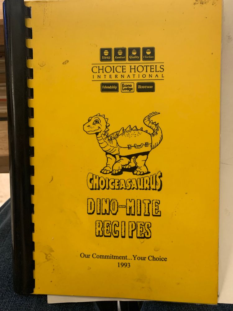 A yellow black plastic bound recipe book. The cover says choice hotels international with a drawing of a dinosaur combine with a suitcase. Underneath the dinosaur it says Choiceasaurus, Dino-mite recipes, followed by our commitment… Your choice 1993