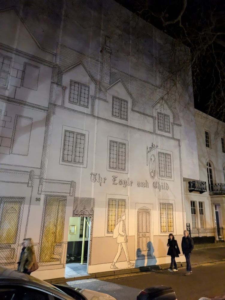 Printed canvas over scaffolding, showing a three-story old building with lead-lined windows and a wooden door. "The Eagle and Child" in lettering across the front. Photo taken at night.