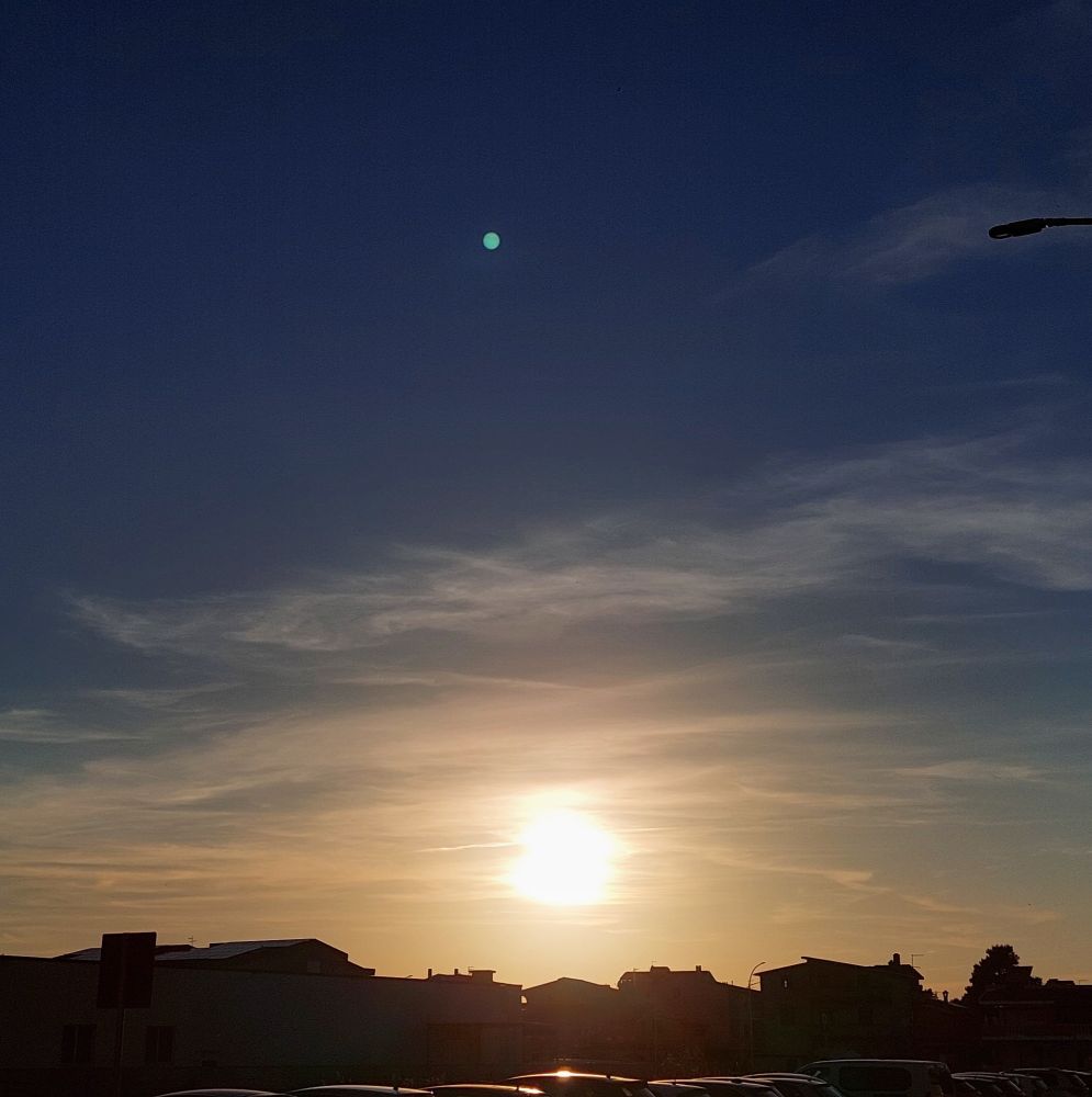 Almost sunset, the sun is visible behind a thin layer of low clouds, like a veil, making them golden. The sky is deep blue. 