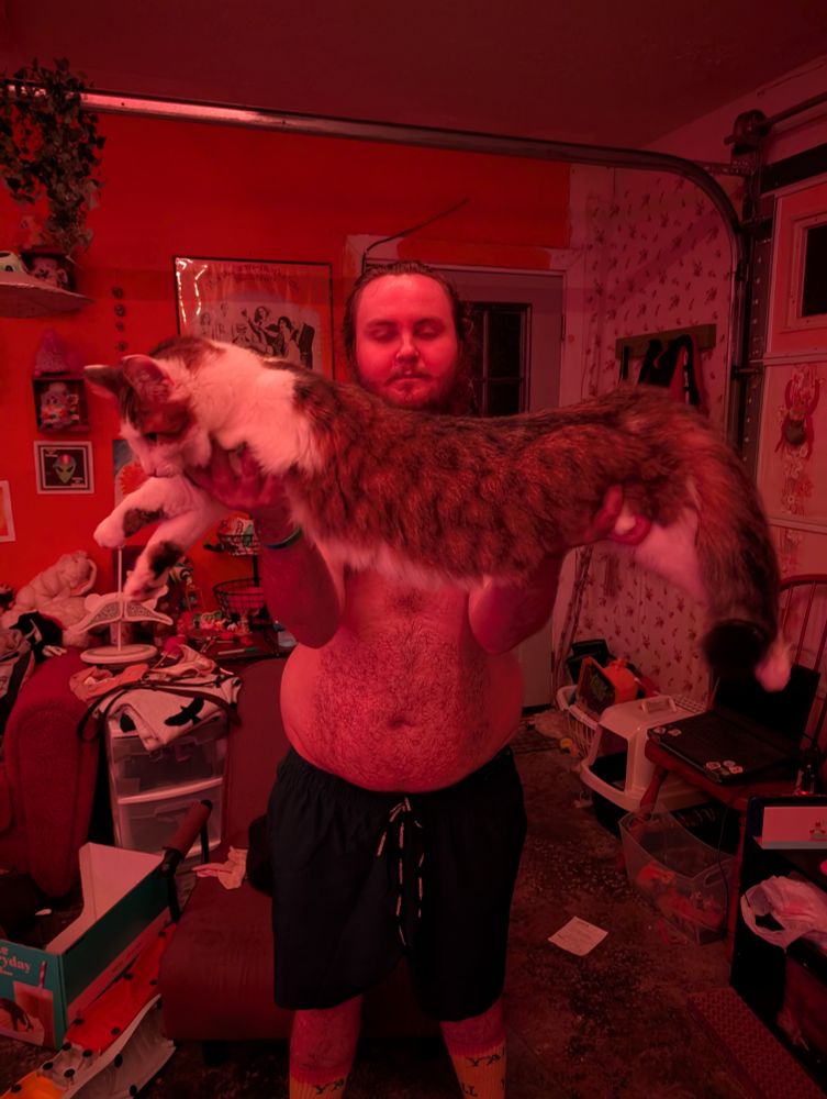 a chubby white nonbinary person, me, holding up my under his waist and armpits. the cat is a long-haired tabby. he is facing the side. 
