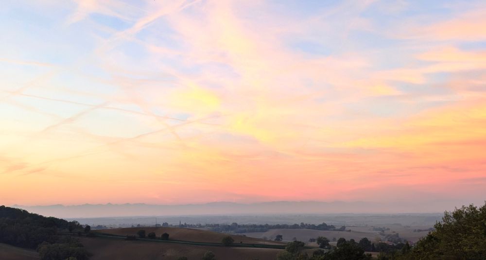 Panorama de collines au soleil levant, ciel en dégradé de couleurs orange, jaune et bleu. Les traces des avions semblent dessiner un pentagramme.