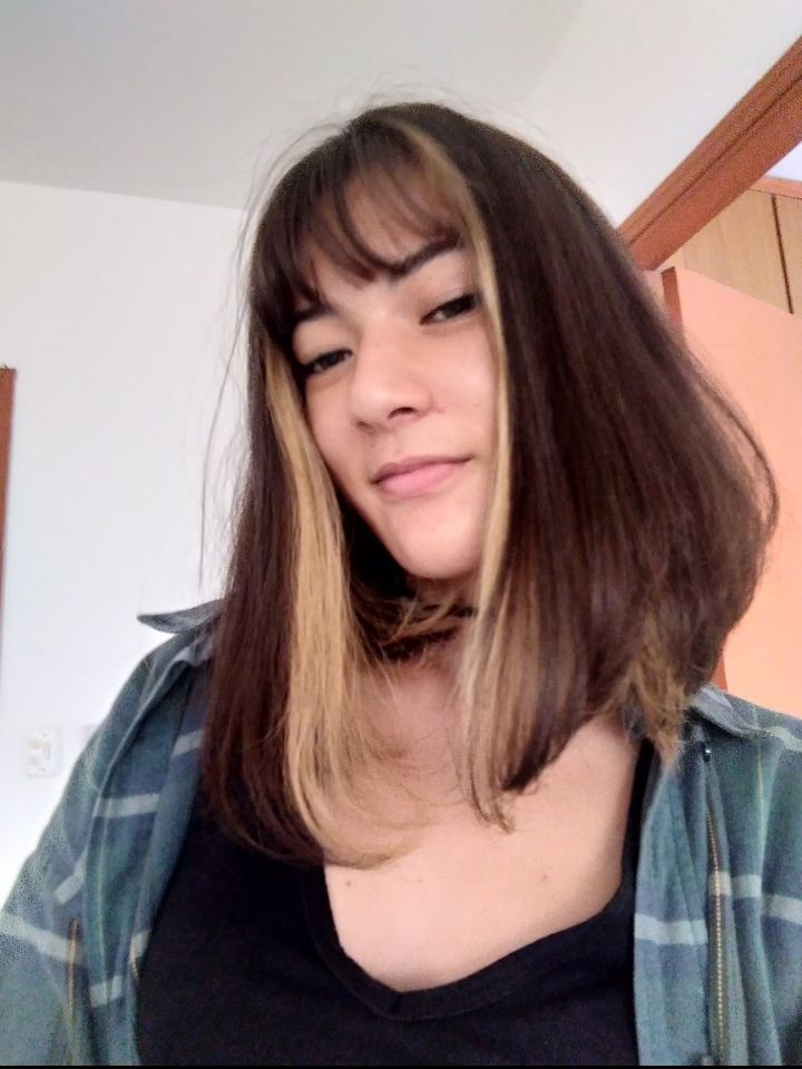 Selfie da dona desta conta em 2019 sorrindo levemente. Meus cabelos castanhos estão lisos, com um corte chanel e com uma mecha loira de cada lado, uso uma camisa xadrez verde por cima de uma camiseta preta.