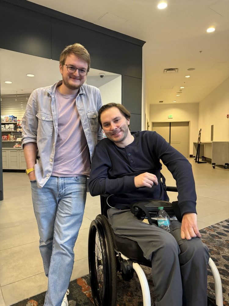 Grian, dark blonde hair and glasses, wearing jeans, a light lilac shirt, and a light gray jacket, standing next to GoodTimesWithScar, brown hair, navy long-sleeved shirt, and gray pants, sitting in a wheelchair with an oxygen tube in his nose. They look very happy.