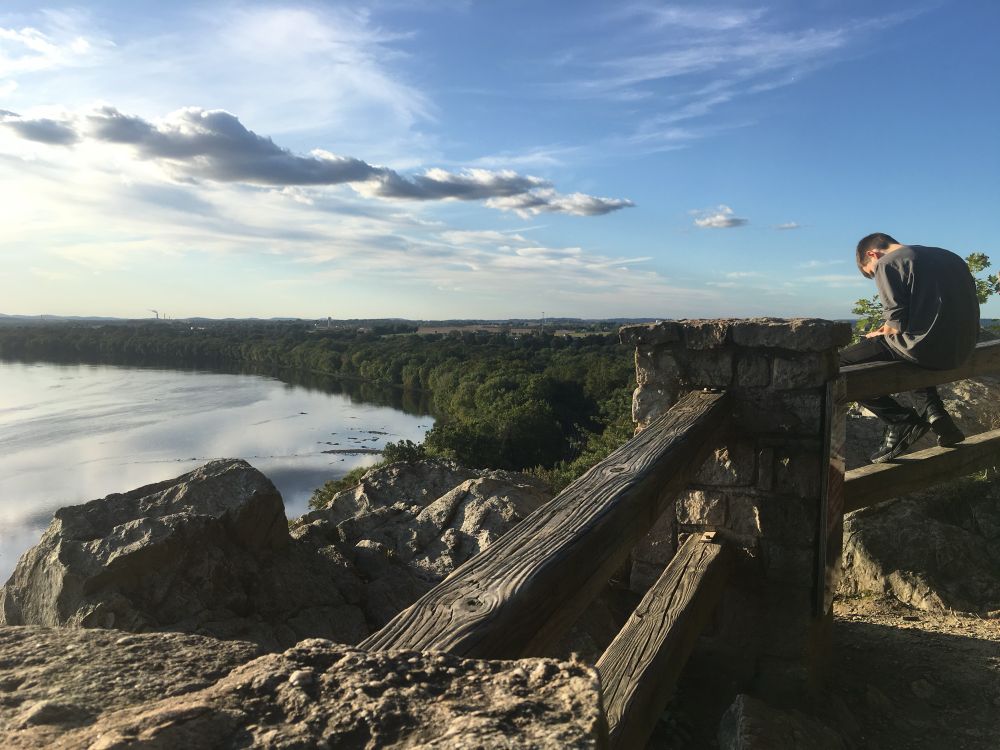 A ridge overlooking a cliff with some woods and a river below, behind a fence with a man sitting on it