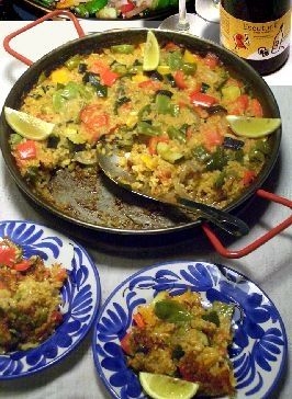 パエリャパンを囲んでの食卓。パエリャパンの中には野菜たっぷりのパエリャがあり、4分の一ほどがお皿に取り分けられている。パエリャの具はトマト、なす、モロッコインゲン、赤パプリカ、黄パプリカ、ズッキーニ、などが見える。櫛形にカットされたレモンが3切れ飾られている。手前に青い花模様の小皿が2つあり、それぞれに取り分けられたパエリャが乗っている。