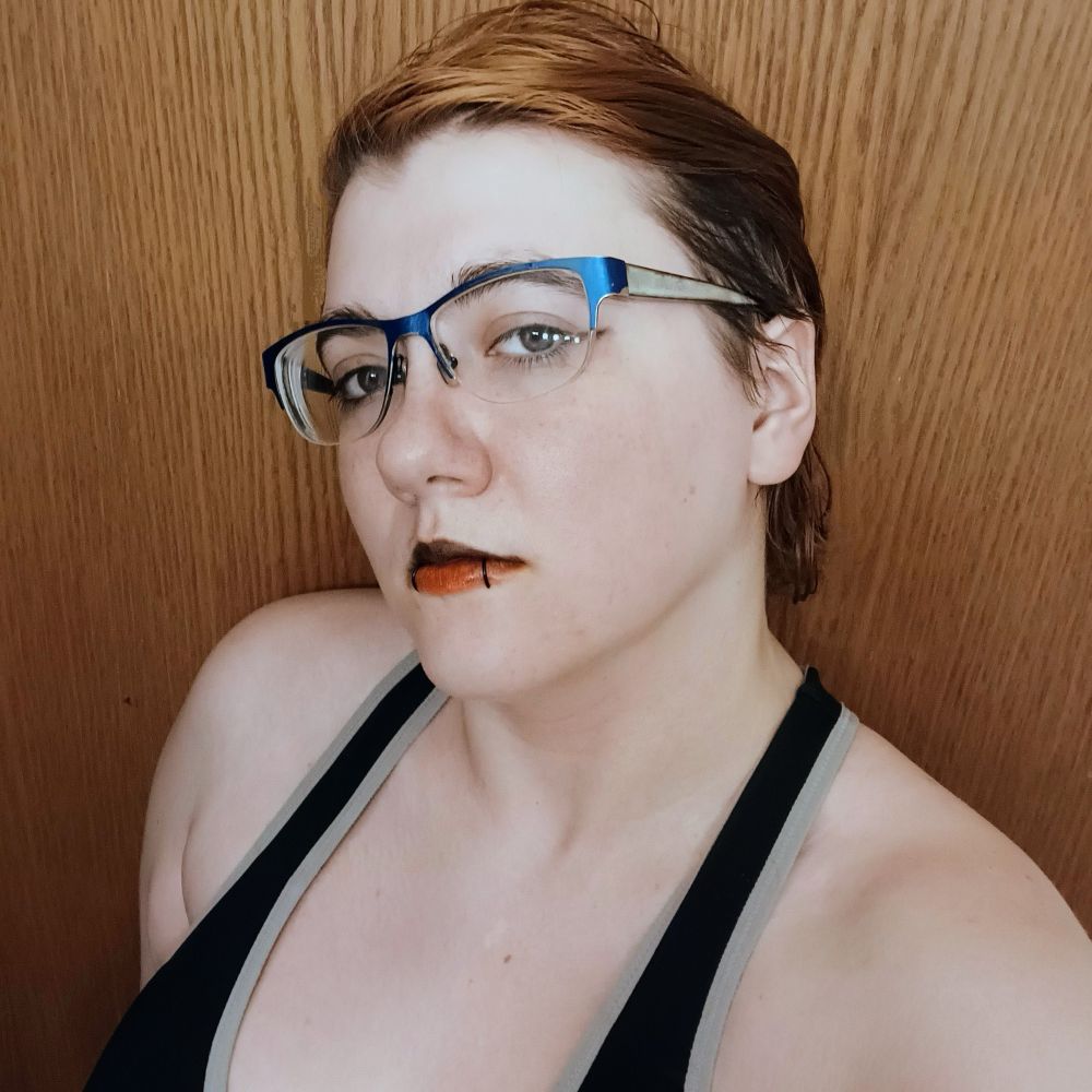 Goddexx Andi with glasses on, slightly hiding the small subtle wing on the eyeliner, the black lipstick on the top lip and a nice reddish orange bottom lip under their snake bite piercings 