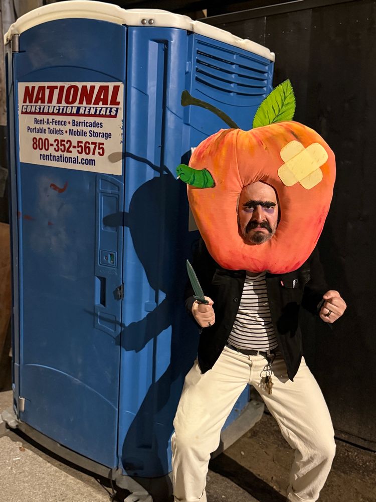 A picture of a man in an apple shaped headpiece outside a portapotty looking menacing 